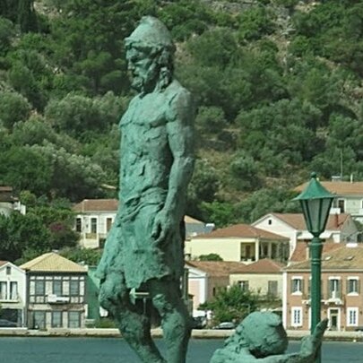 A green statue of Odysseus in Greece with buildings at the base of a mountain of greenery. 