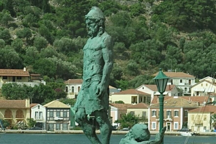 A green statue of Odysseus in Greece with buildings at the base of a mountain of greenery. 