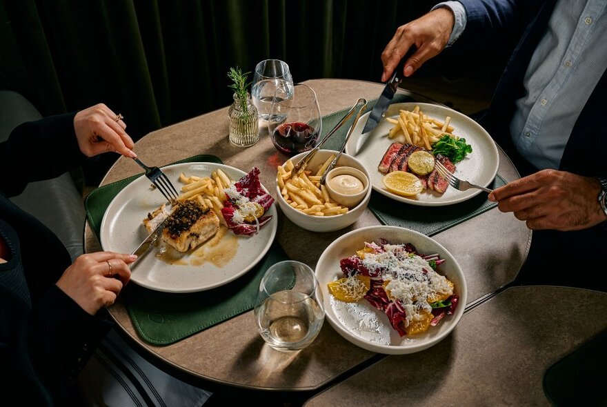 A couple seated at a restaurant table with knives and forks and plates of food with chips, drinks and sides.