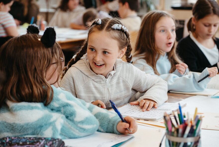 Primary school aged children seated at a large table writing and interacting during a creative writing workshop in a bright studio space.