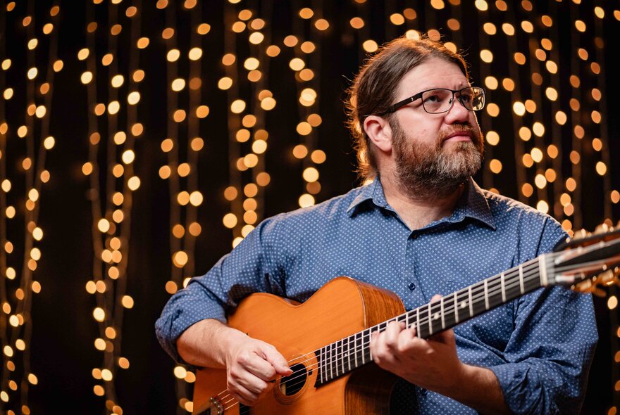 Bearded guitarist wearing blue shirt and spectacles, rows of lights behind him.