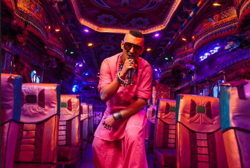 Pakistani rapper Faris Shafi holding a microphone close to his face and performing in the centre aisle of a space that has rows of airline or bus seats on either side of him. 