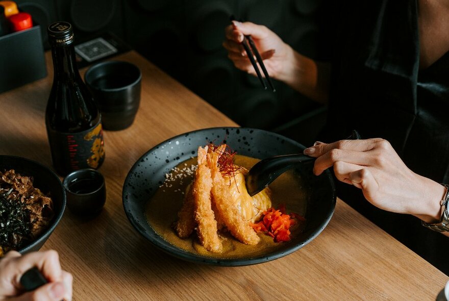 Person placing a spoon and chopsticks into a bowl of food at a table.