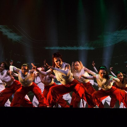 A large group of dancers from the Royal Family Dance Crew, in white tops and red pants perform a high-energy routine on a dark stage with dramatic lighting.