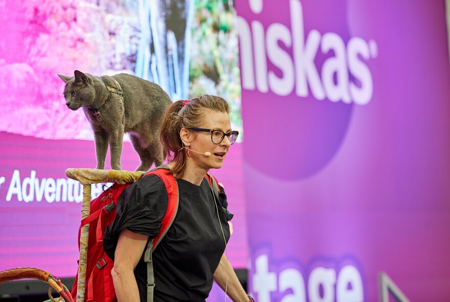 A cat on the back of a woman's shoulders at an expo with a Whiskas purple backdrop.