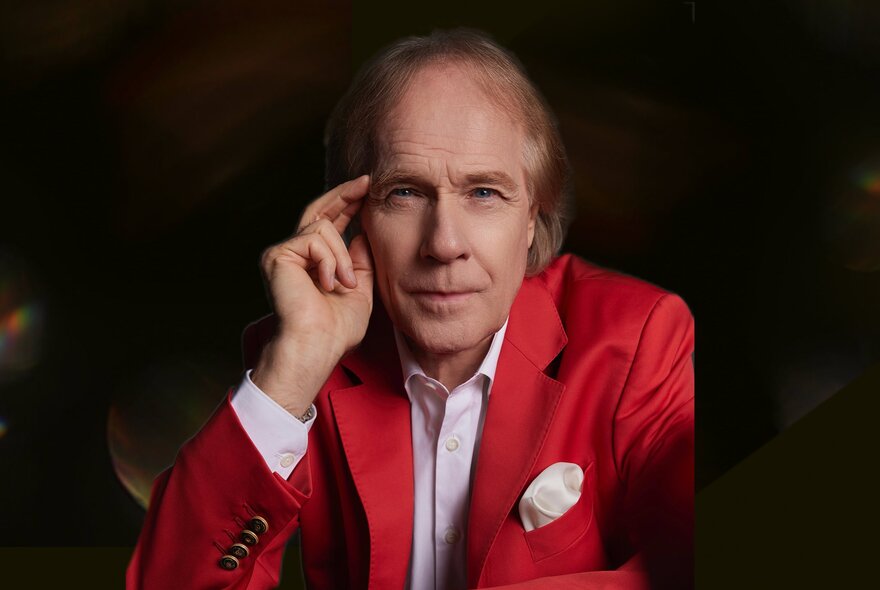 Pianist Richard Clayderman wearing a red jacket and white shirt, he holds his hand near his face and is posed against a dark background.
