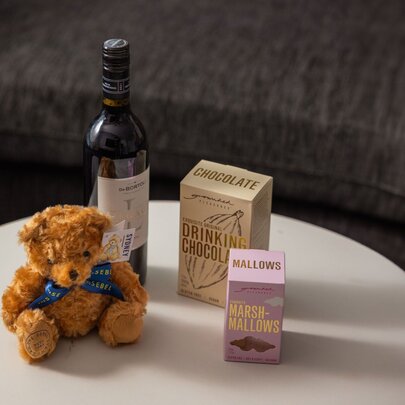A small teddy bear soft toy, a bottle of wine and two boxes of premium drinking chocolate resting on a round white table.