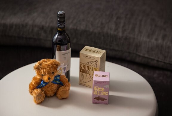 A small teddy bear soft toy, a bottle of wine and two boxes of premium drinking chocolate resting on a round white table.