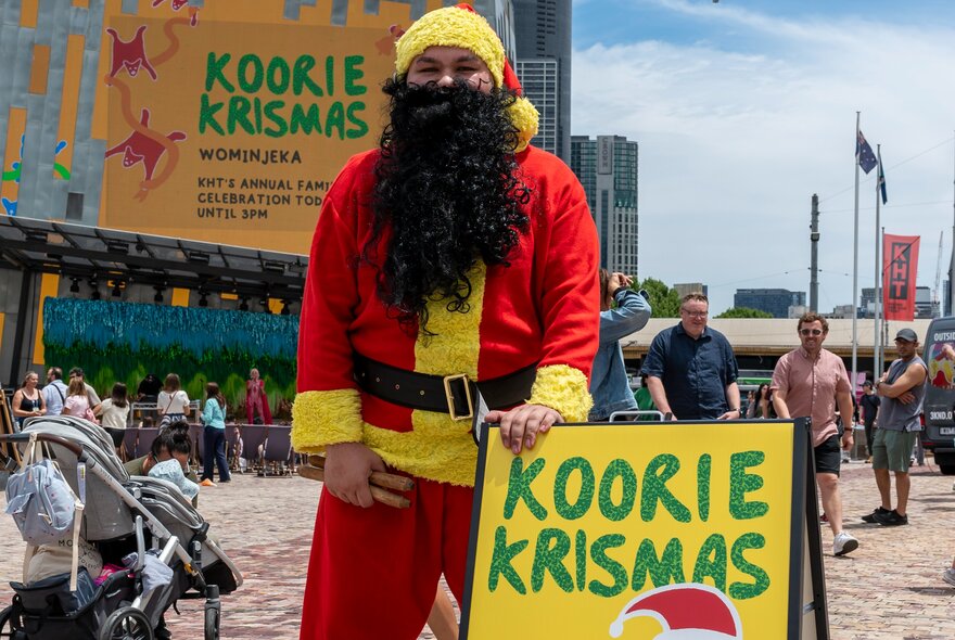 Koorie Klaus, wearing a red and yellow suit with a black beard, standing outside at Fed Square, next to a sandwich board sign with the words KOORIE KRISMAS written on it.