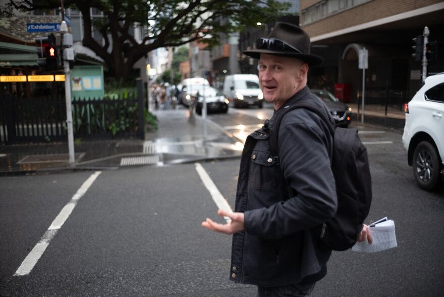 A man wearing a black hat and talking, leading a walking tour across a Melbourne city street.