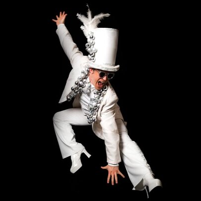 A man dressed as Elton John with a white suit and comically tall top hat and high-heeled boots, stretching in a flamboyant pose. 