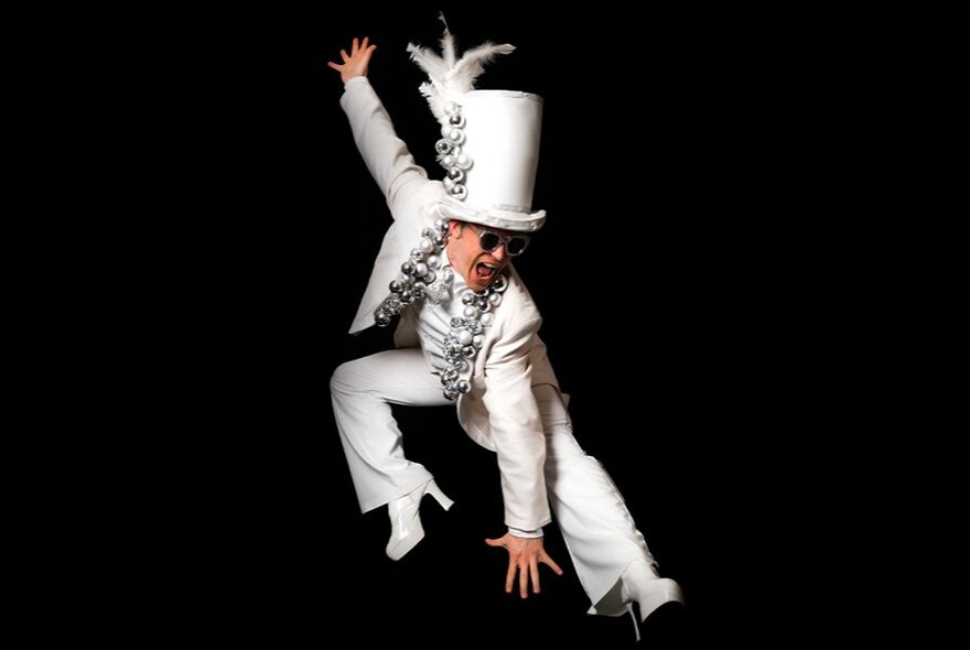 A man dressed as Elton John with a white suit and comically tall top hat and high-heeled boots, stretching in a flamboyant pose. 