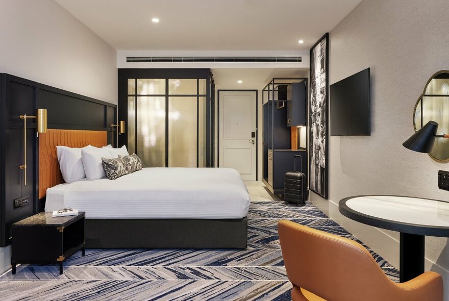 A hotel room with white king size bed, pillows, patterned carpet, and a small table, lamp and armchair in the foreground.