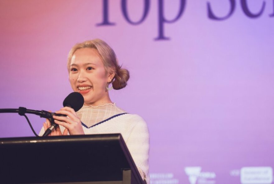 A young woman standing at a podium holding a microphone, in front of a purple digital backdrop.