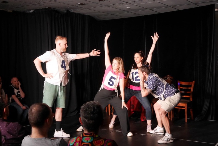 An ensemble cast of impro actors on a stage, performing in front of a live audience in a small theatre.