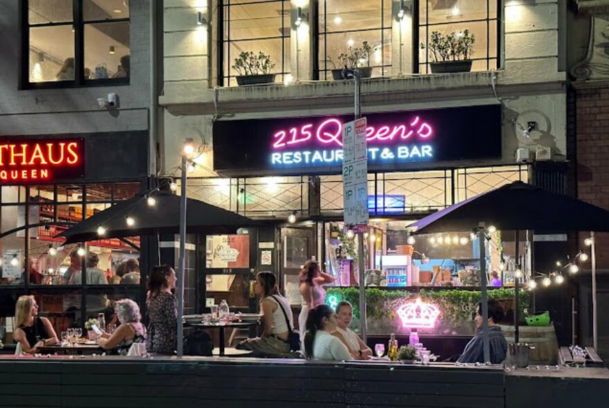 Street scene showing outdoor diners seated in front of a restaurant, marquees, fairy lights and neon signs also visible.