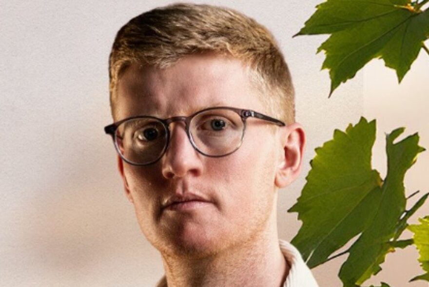 A man with neat hear and glasses looks serious in front of a wall with plant leaves to the right of the image. 