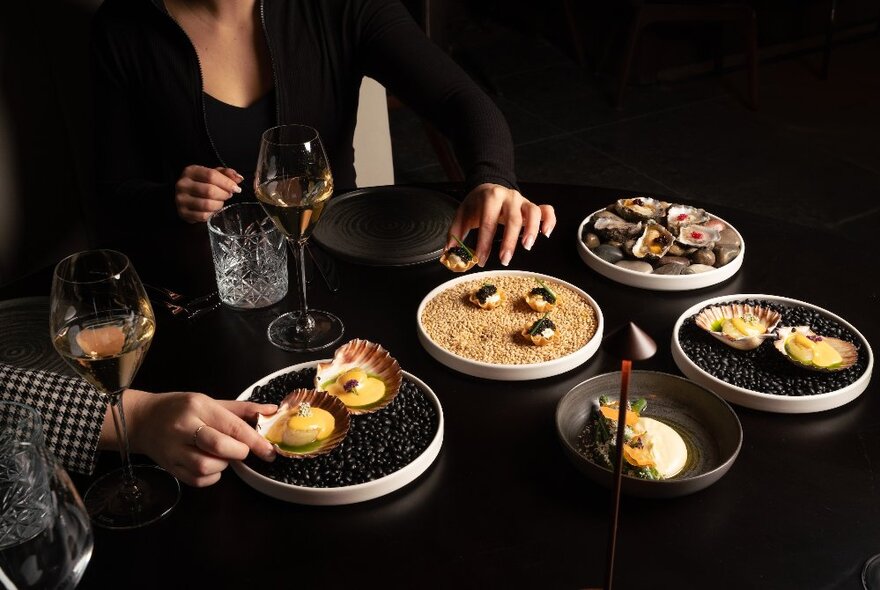 People taking food from round white dishes of food on a black table with glasses of wine.