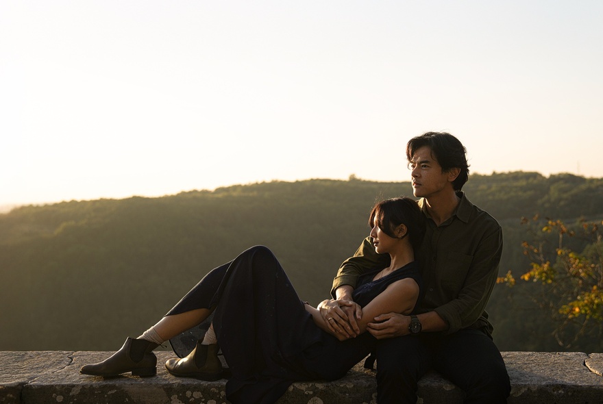 A still from the movie SORE: Wife From the Future with a woman leaning back into a man's arms as they sit on a wall with hills in the background.