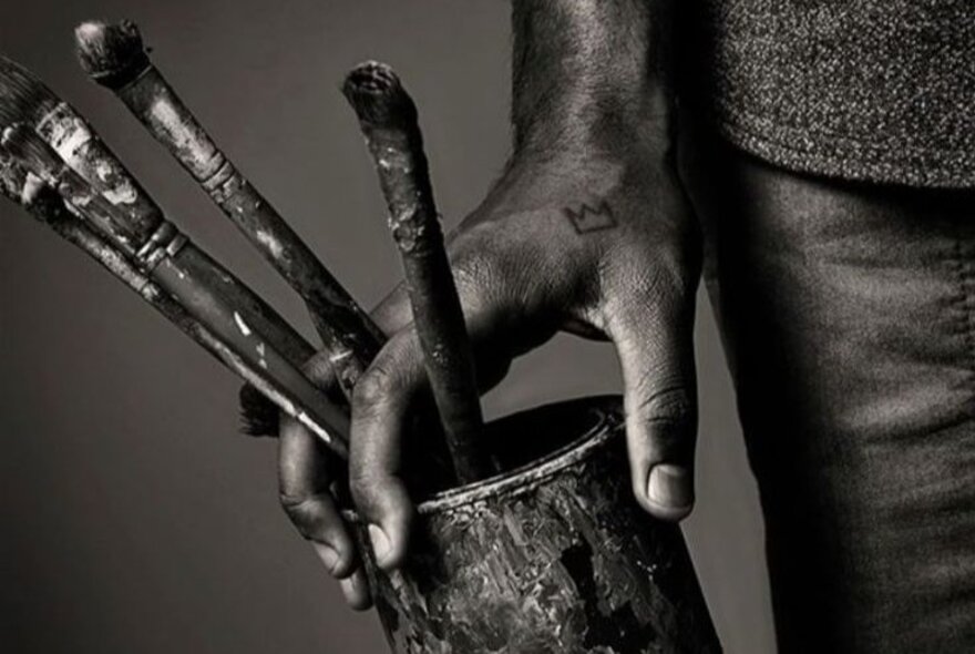 A black and white photograph of a person's hand holding a paint-covered cup with four paint brushes sticking out of it.