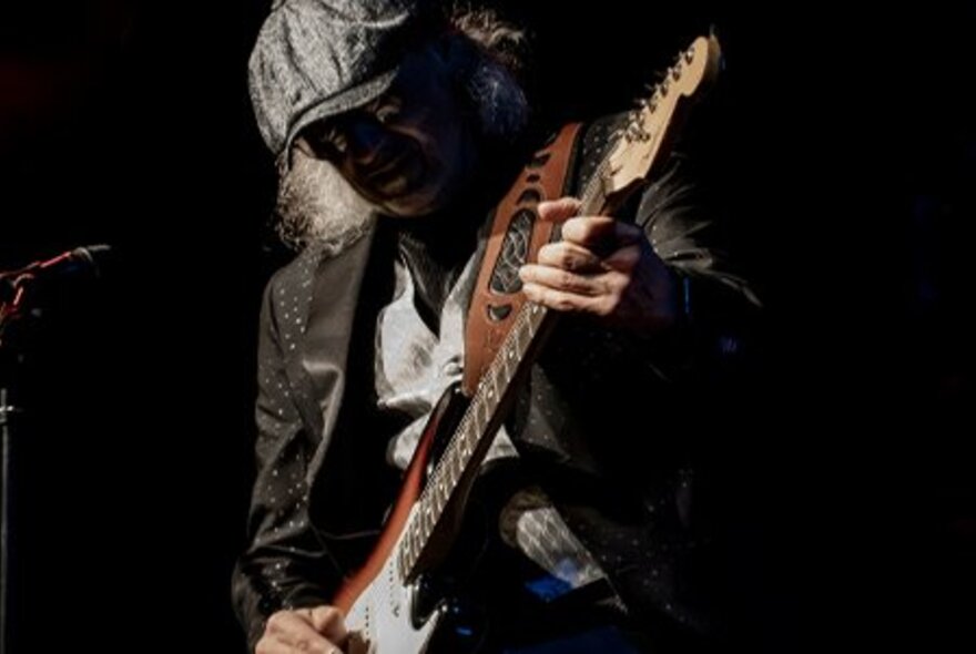 Man wearing a peaked cap that casts a shadow on his face, playing lead guitar on stage.