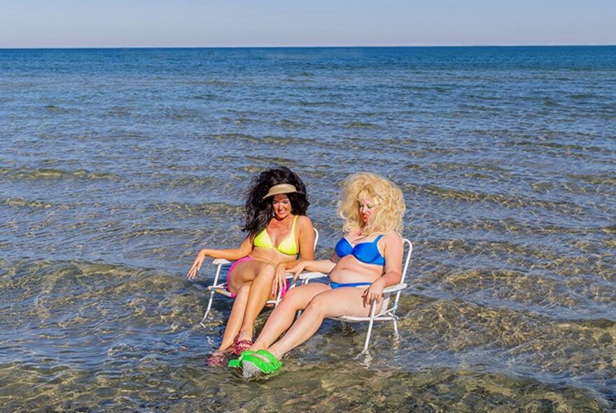 The two performers from the show BIRDS, both wearing bikinis and with big hair, seated in beach chairs in the shallow water at the beach.