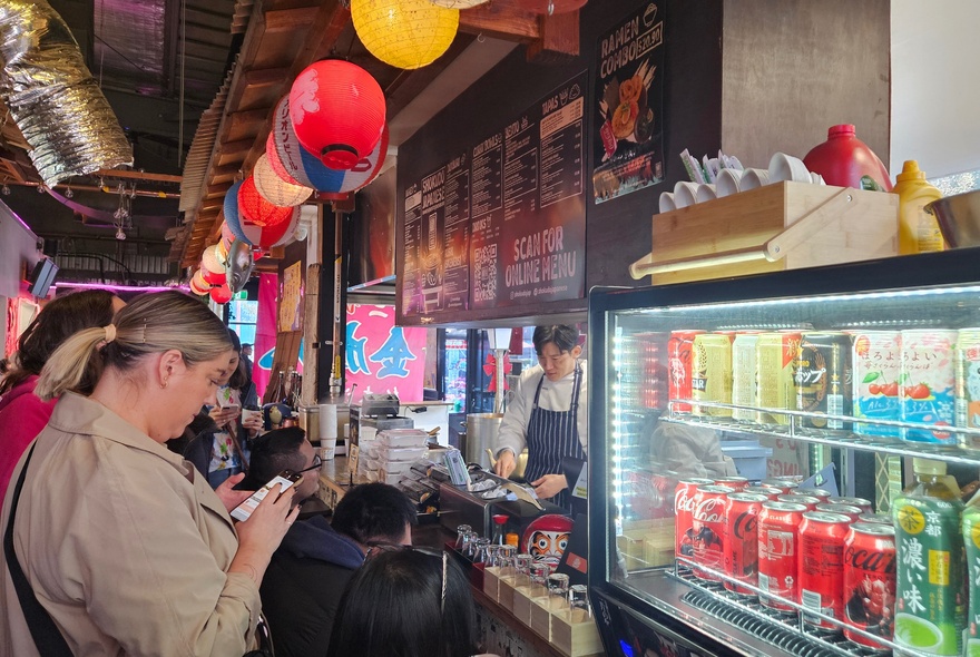A person looking at her phone, standing in front of a drinks fridge filled with brightly coloured cans, inside a bustling food night market.