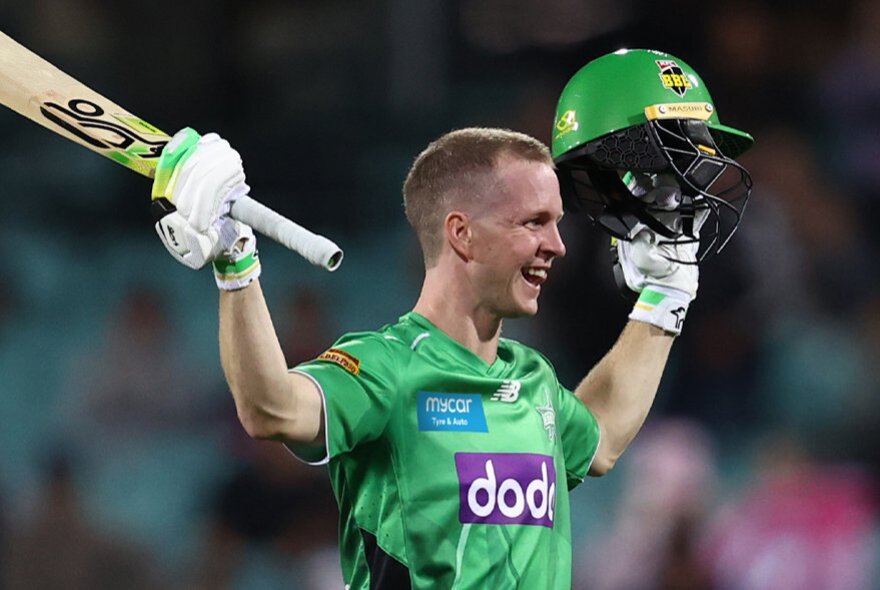  A smiling male cricketer in a bright green Melbourne Stars jersey celebrates a milestone by raising his cricket bat in his right hand and holding up his green helmet in his left hand.