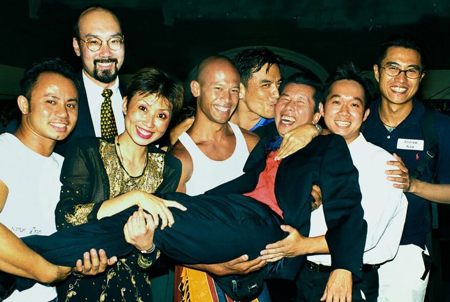 A large group of friends all smiling and holding up a central person horizontally, one kissing him in the head as he laughs.