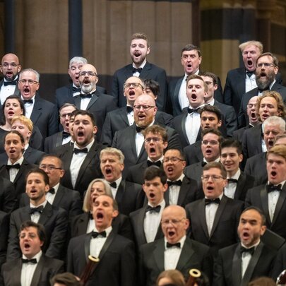 A large choral ensemble performance, with the singers wearing black clothing, all of them singing.