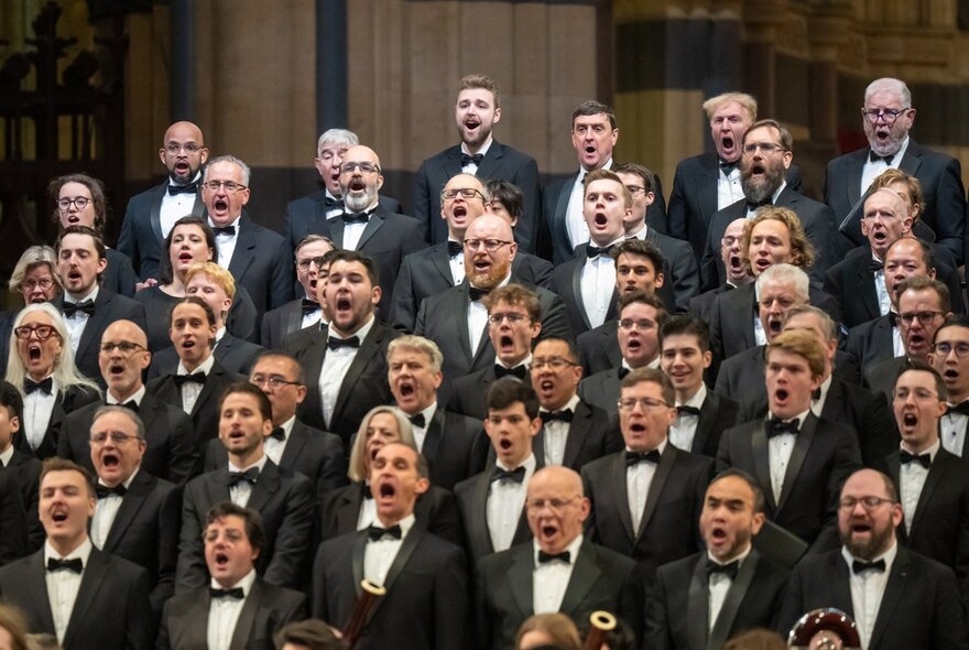 A large choral ensemble performance, with the singers wearing black clothing, all of them singing.