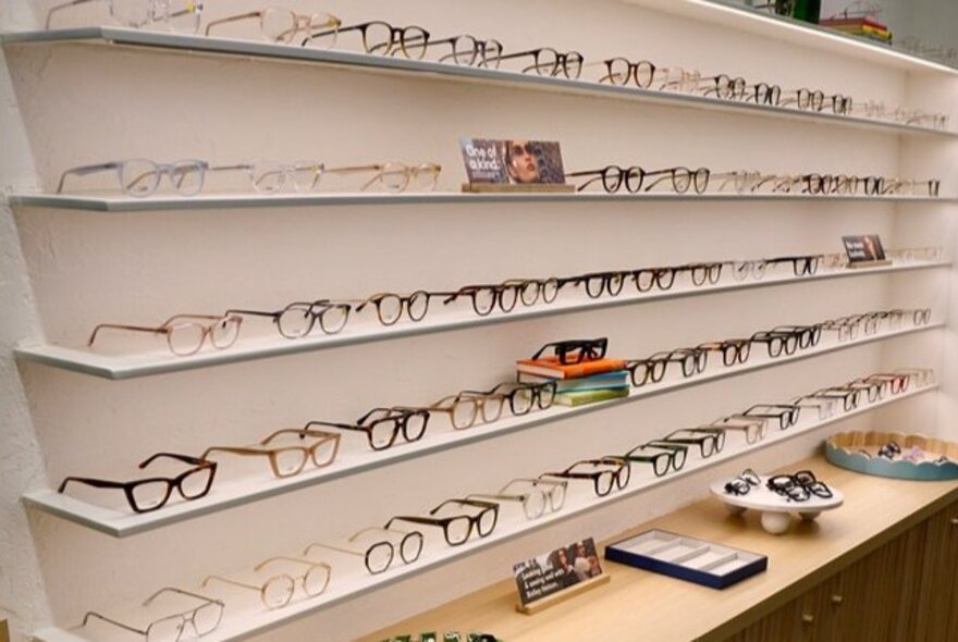 Rows of glasses displayed on floating shelves on the white wall of an eyeglass store.