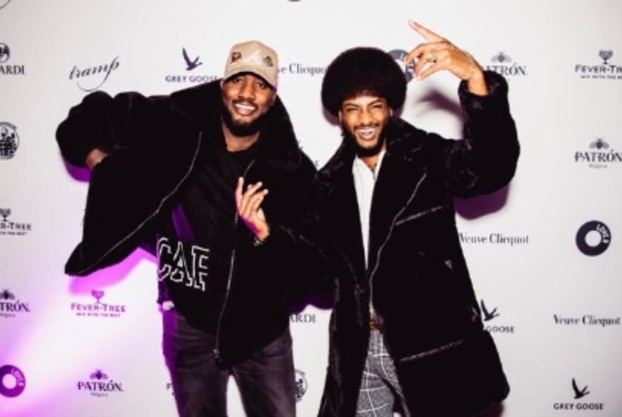 Two smiling men, in heavy fur coats, posing against a sponsor wall.