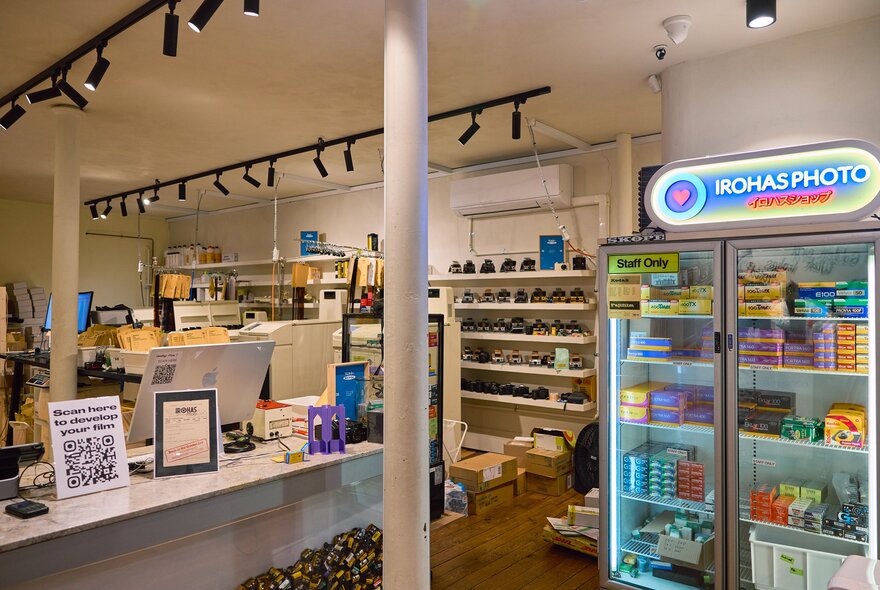 Inside a film developing shop there is a glass cabinet filled with film and above it is a neon sign saying irohas photo