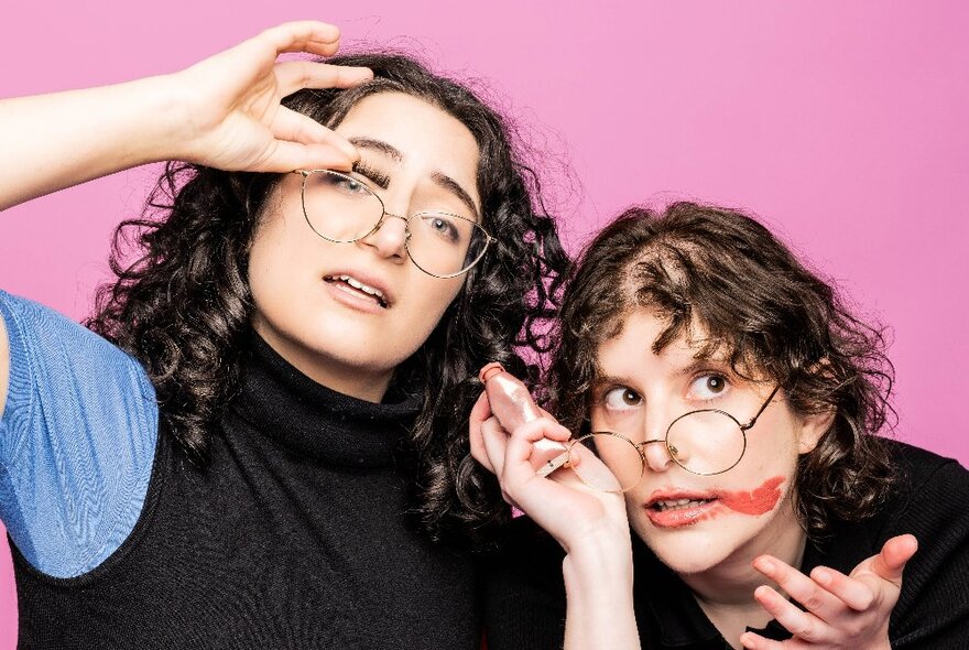 Two comedians close to each other, one with smeared lipstick and the other lifting her glasses off her nose - both looking slightly panicked and messy. 