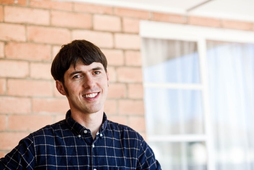 Portrait of a smiling young man in a blue checked shirt, with a residential brick wall and windows behind him.