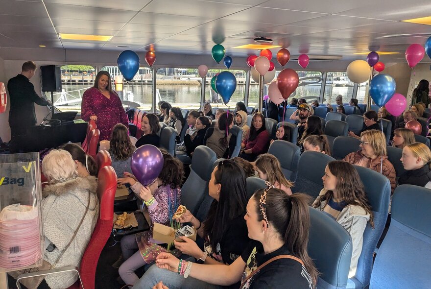 Seated children with balloons in a low-roofed cabin, looking at a performer and DJ.