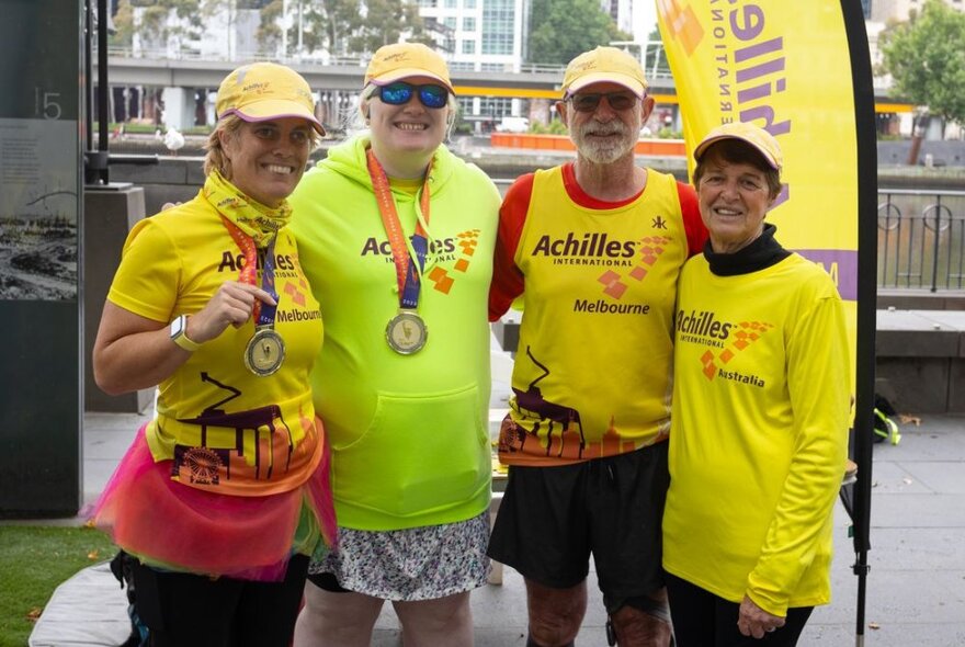 Four people lined up and posing for a photo, all wearing fluoro yellow tops; two of them holding medals they wear around their necks, at an outdoor festival.