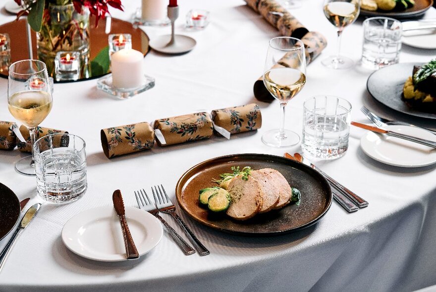 A table laid with white linen, plates of food, cutlery, glasses of wine and Christmas decorations including candles and Christmas bon bon crackers.