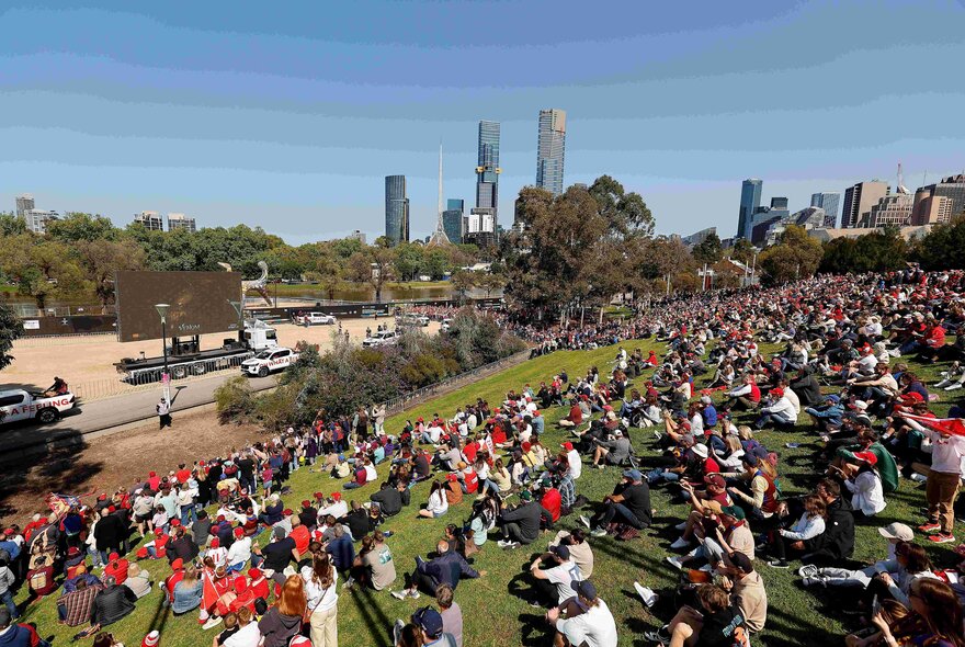 Crowds of people seated on the grassy low slopes of Birrarung Marr in anticipation of the Grand Final Parade motorcade passing by.