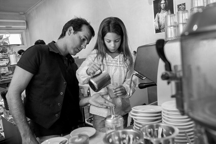 Man watching a child pouring milk into a coffee drink with expresso machine and cups.