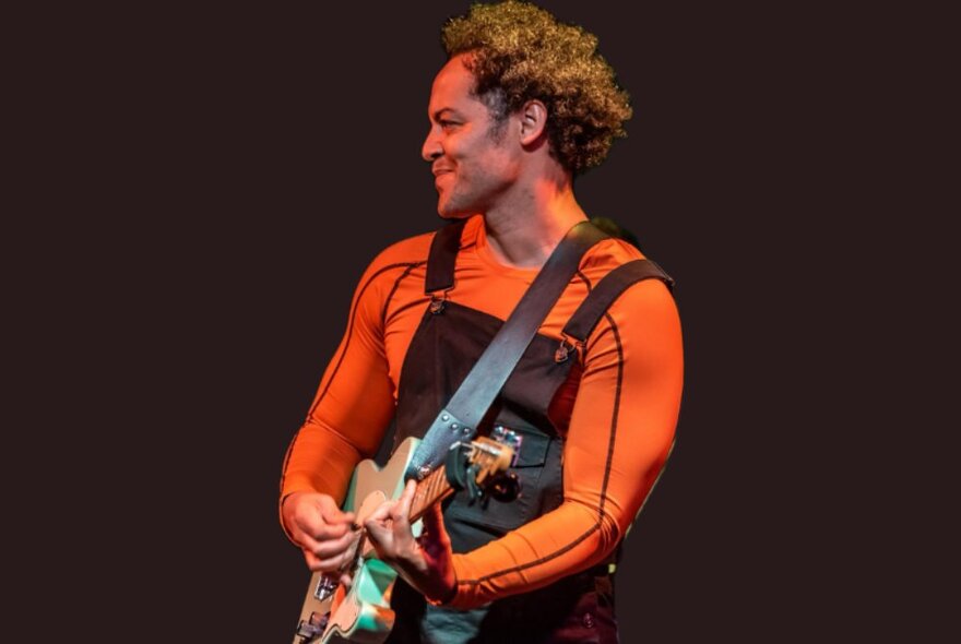 A man wearing an orange long sleeved top and playing an electric guitar on stage, in profile view.