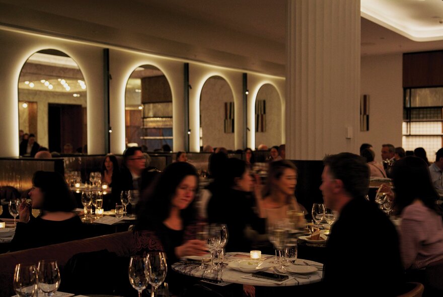 A dimly lit dining space with people eating at tables and archways lit in the background. 