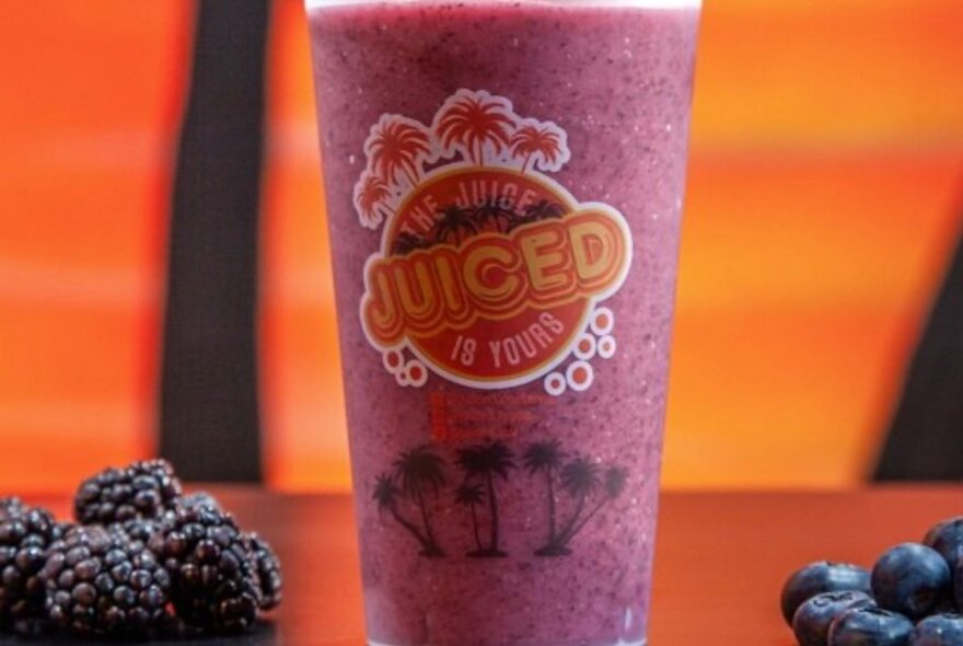 A takeaway plastic cup of purple coloured juice, with branding on the cup, blackberries and blueberries next to it.