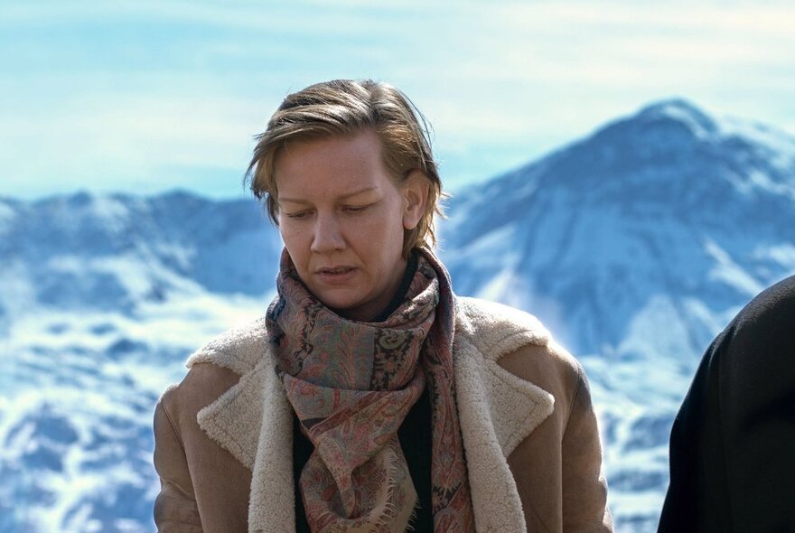 Woman wearing winter clothes standing in a snowy alpine setting, looking down.