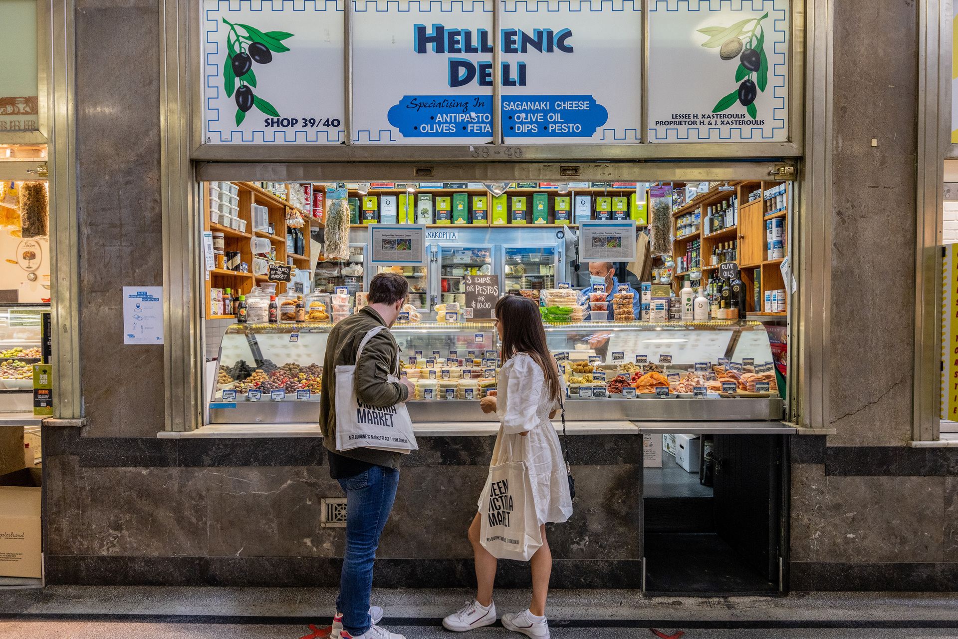 Two friends are looking at deli goods at a Greek shop in a market 