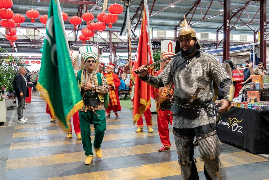 A group of men wearing red and green cloaks, some bearing large flags, in an open space at Queen Victoria Market, with a crowd of onlookers watching.