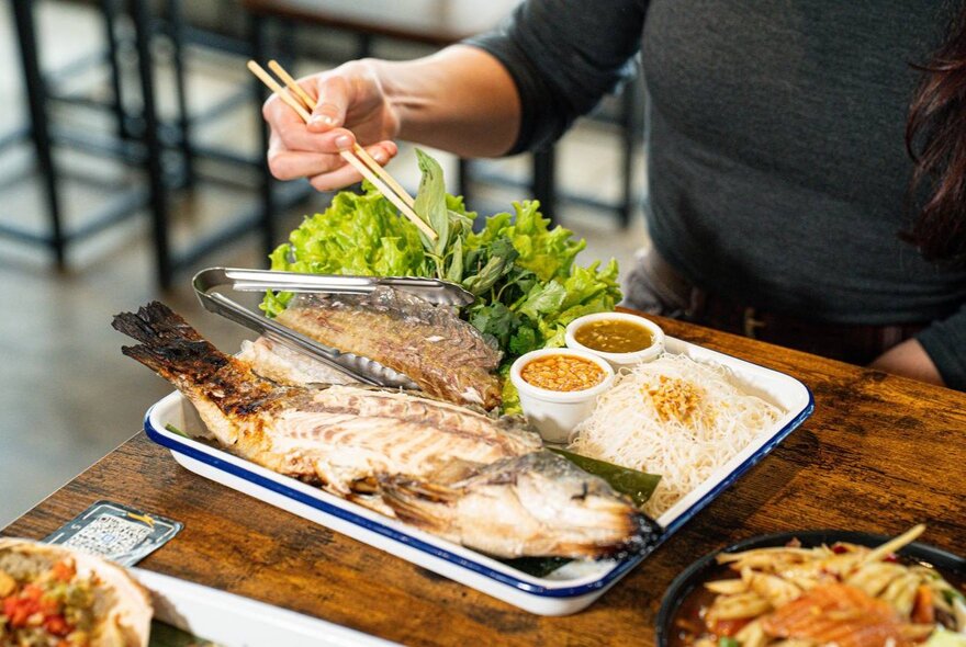 Hand holding chopsticks over a plate of fish with rice and garnishes.