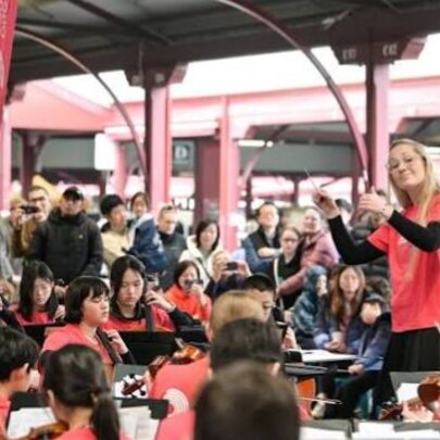 The Melbourne Youth Orchestras performing in an outdoor undercover space at the Queen Victoria Market, a conductor leading them, and an audience standing around watching.