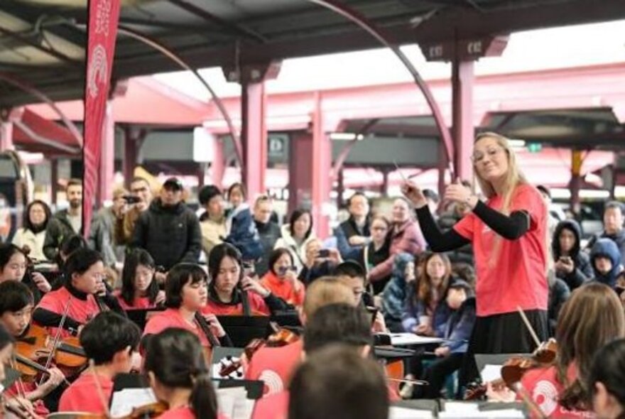 The Melbourne Youth Orchestras performing in an outdoor undercover space at the Queen Victoria Market, a conductor leading them, and an audience standing around watching.